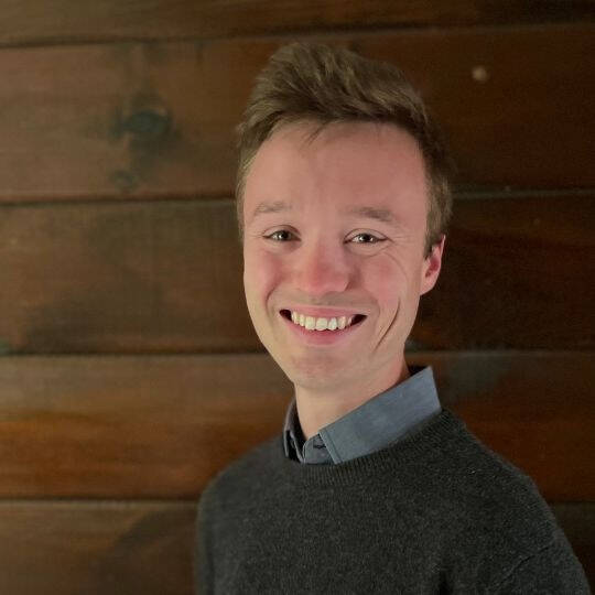 An individual smiles at a camera wearing a gray shirt and cover sweater in front of woody background